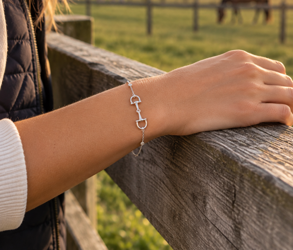 A person rests their hand on a wooden fence, wearing a silver horse bit bracelet. In the background, horses graze in a green paddock at sunset.