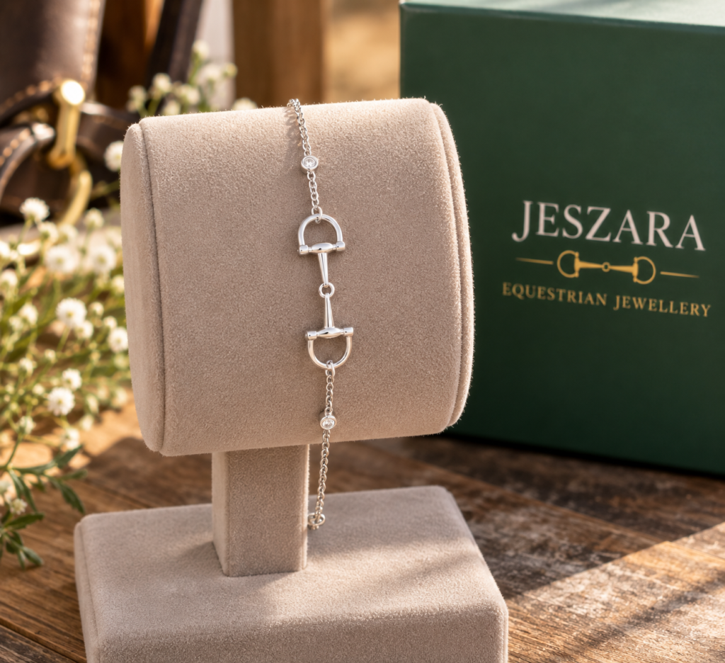 A silver equestrian-themed bracelet displayed on a beige stand, with a green JESZARA Equestrian Jewellery box and small white flowers in the background.