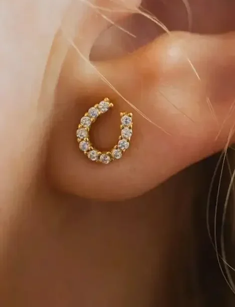 Close-up of an ear adorned with the Luxe Horseshoe Earrings—gold, horseshoe-shaped, and encrusted with small clear gemstones. The earring is attached to the earlobe, with strands of blonde hair visible at the edge.