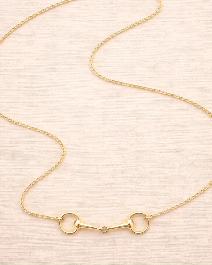 The Snaffle Bit Necklace features a delicate gold chain adorned with a horse bit-shaped pendant, elegantly displayed on a light beige textured surface—making it the perfect piece of equestrian jewellery for any horse lover.