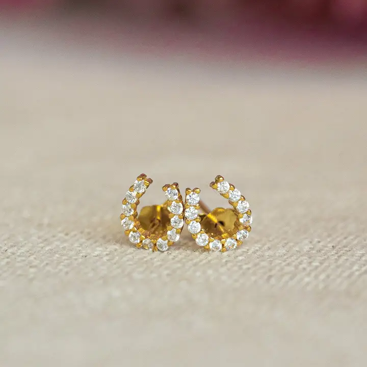 The Luxe Horseshoe Earrings are a pair of small, gold studs shaped like horseshoes and adorned with sparkling white stones. Displayed on a neutral textured surface, these elegant earrings offer a timeless addition to any luxury jewellery collection.