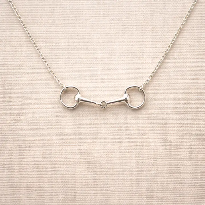 The Snaffle Bit Necklace is an elegant piece of equestrian jewellery, crafted in delicate silver with a horse bit–shaped pendant, displayed against a neutral beige fabric background—perfect for horse lovers.