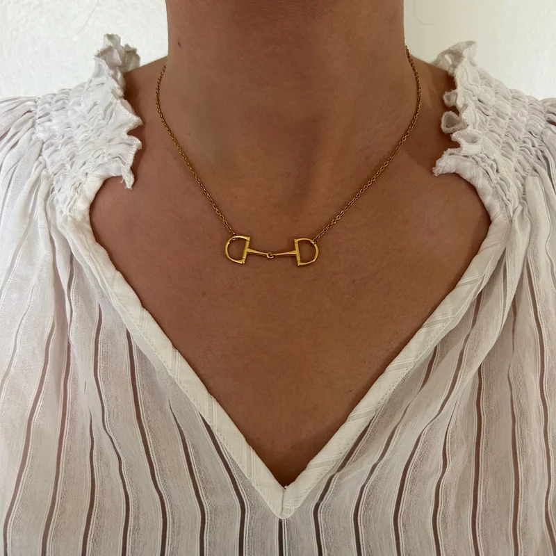 Wearing the Oly Necklace, a gold accessory featuring a horse bit design, paired with a white blouse detailed with ruffled edges and vertical stripes.