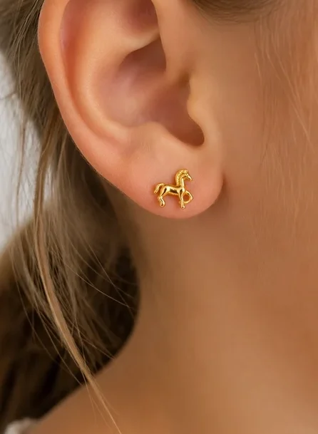 A close-up highlights a young person’s ear adorned with Tiny Horse Earrings—delicate gold horse-shaped studs. Brown hair is tucked behind the ear, and light skin is visible against a softly blurred background.