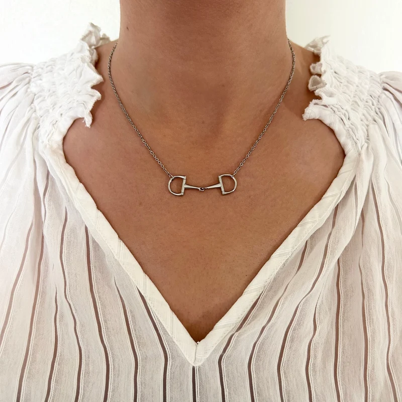 A person wearing a white, pleated, V-neck blouse accessorised with the Oly Necklace, which features a silver horse bit design.