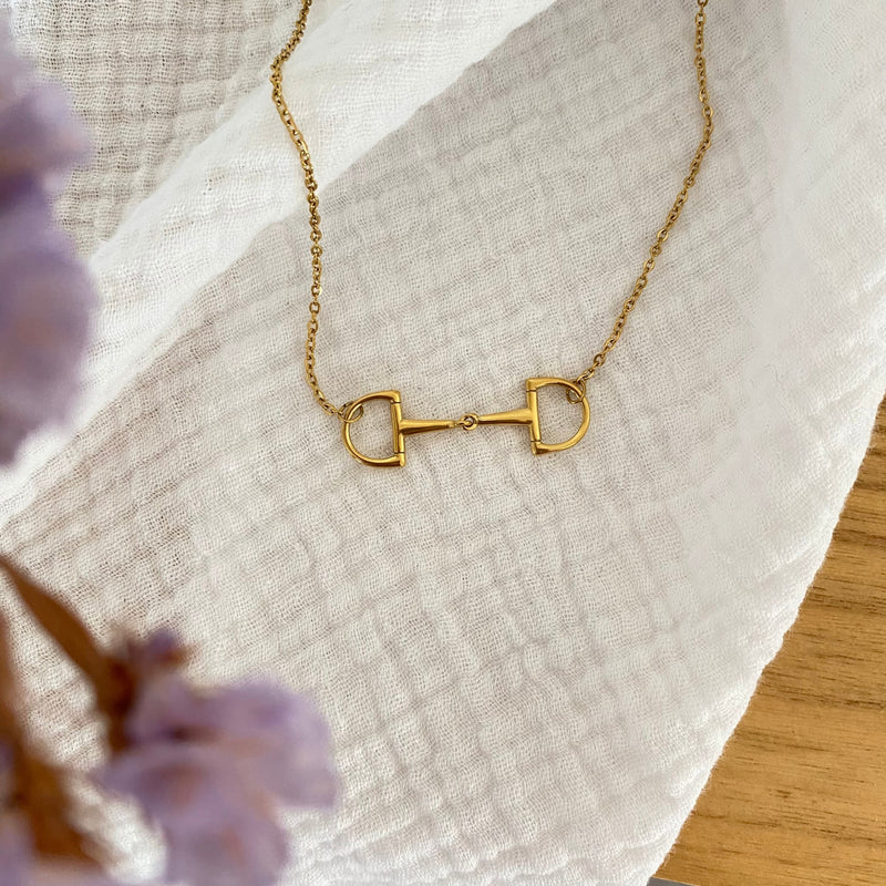 The Oly Necklace, adorned with a gold horse bit pendant, is elegantly displayed on a textured white cloth, accented by softly blurred lavender flowers in the foreground and hints of a wooden surface in the background.