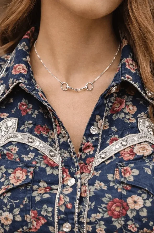 A woman wearing a blue floral western shirt with decorative stitching and pearl snaps, accessorised with the elegant Snaffle Bit Necklace—a perfect piece of equestrian jewellery.