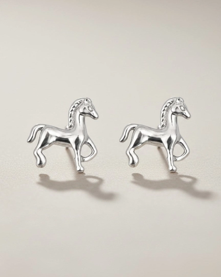 The Tiny Horse Earrings, crafted in silver and delicately shaped like horses, are showcased on a neutral background with gentle shadows beneath each earring.