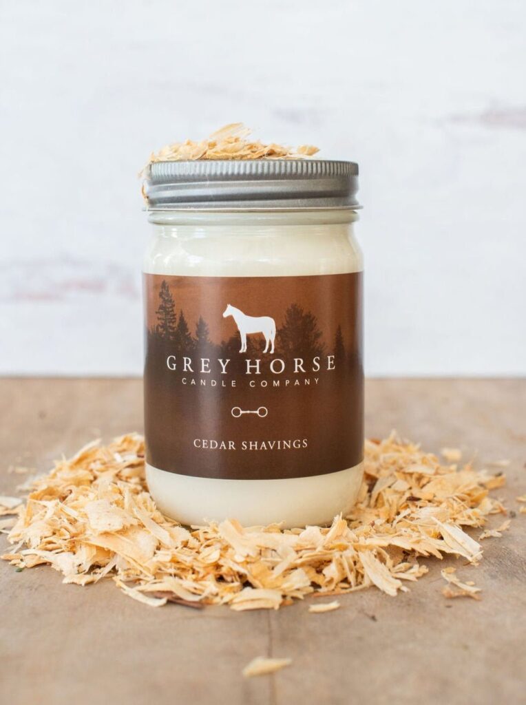 A Grey Horse Candle Company jar labelled Cedar Shavings sits on a wooden surface surrounded by cedar shavings, with a horse silhouette and forest on the label.