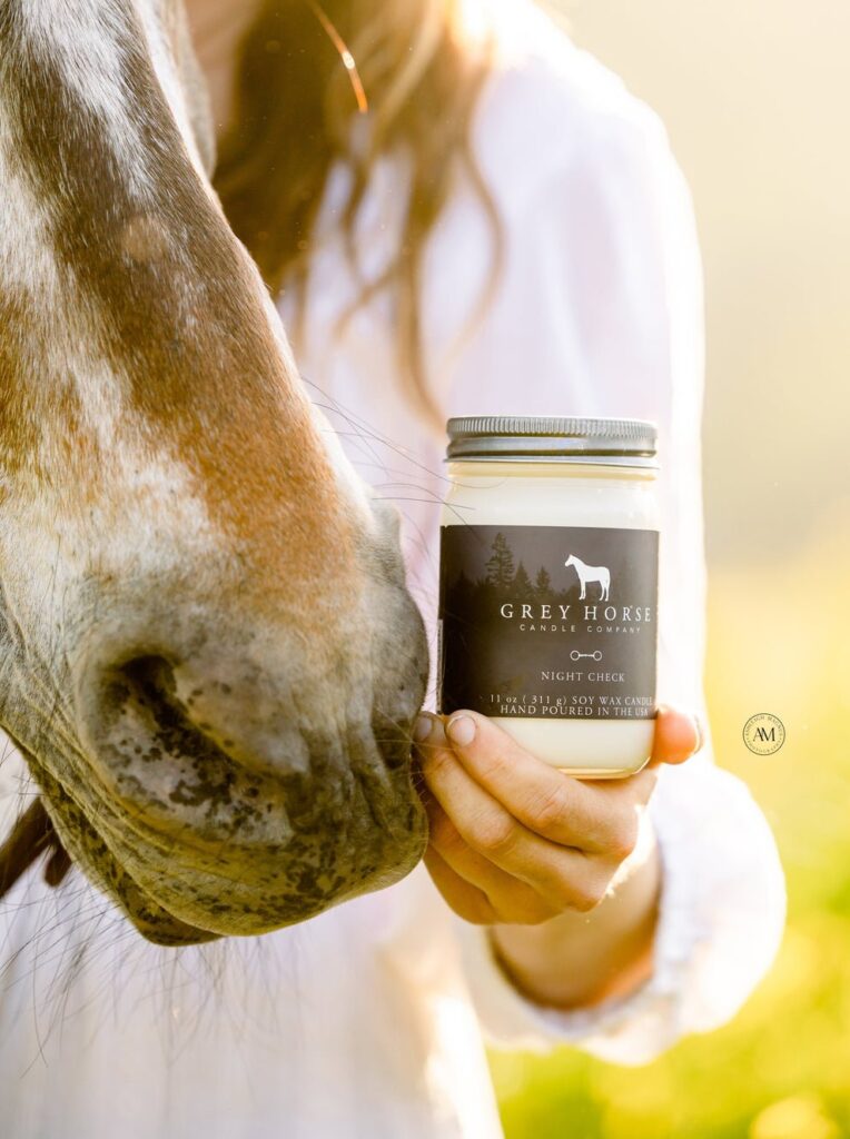A woman in a white shirt holds a Grey Horse Candle Co. jar near a horse's nose outdoors in warm sunlight.
