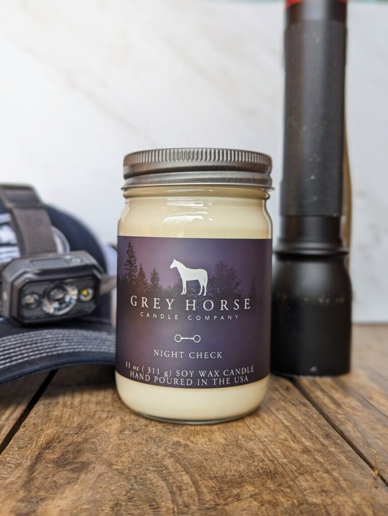 A Grey Horse Candle Company soy wax candle labelled Night Check sits on a wooden surface next to a torch and a head torch on a cap. The candle jar features a horse silhouette and trees on its dark label.