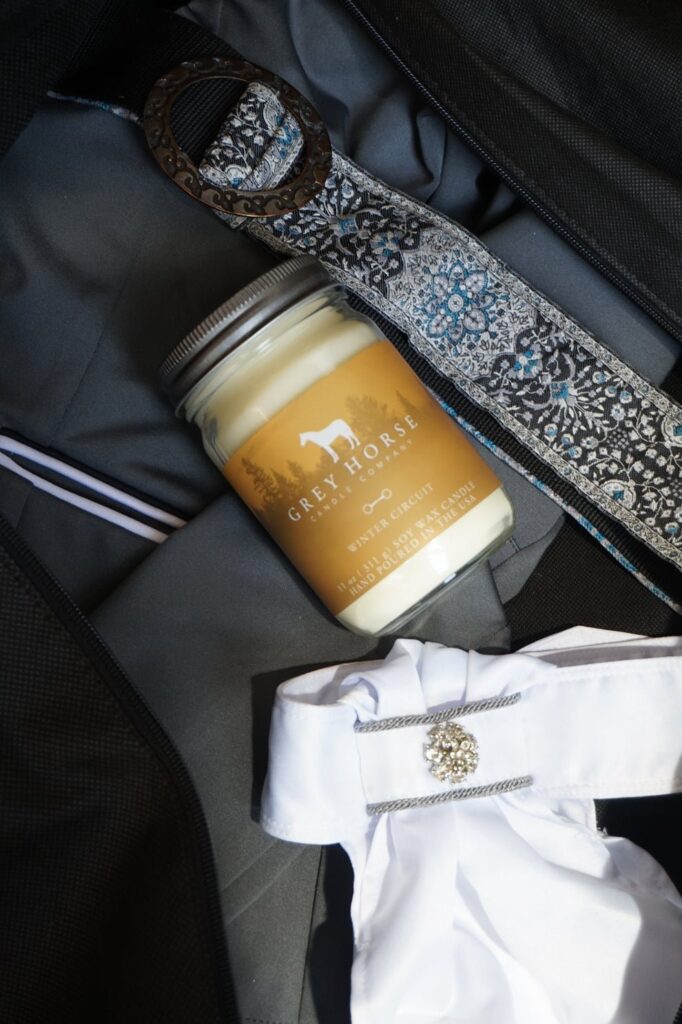 A Grey Horse soy candle, an ornate belt, and a white shirt with a decorative pin are arranged on a dark fabric background.
