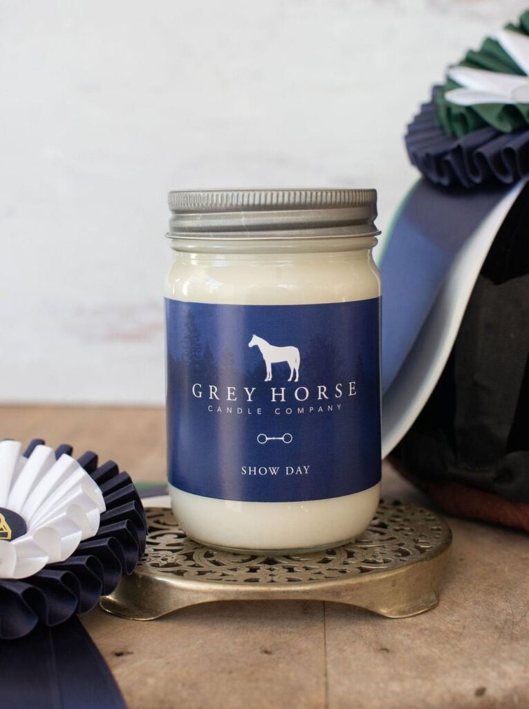 A white candle labelled Grey Horse Candle Company—Show Day sits on a round metal stand, next to blue and white award rosettes and a dark riding helmet.