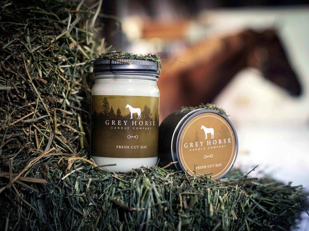 Two Grey Horse Candle Company Fresh Cut Hay candles, one in a jar and one in a tin, sit on a pile of hay. A blurred brown horse is visible in the background.