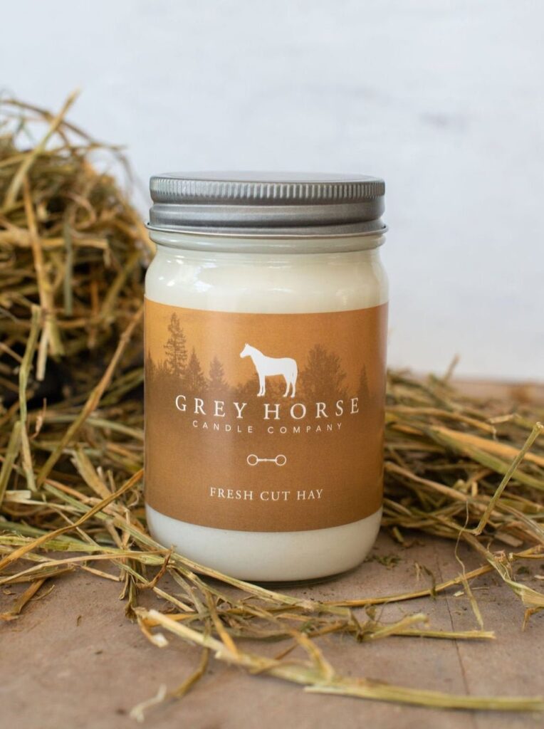 A white candle in a glass jar labelled Grey Horse Candle Company - Fresh Cut Hay sits on a wooden surface with hay in the background.