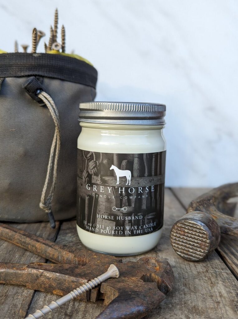 A Grey Horse Candle Company jar candle labelled Horse Husband sits on a wooden surface, surrounded by a hammer, spanner, screws, and a grey tool bag. The background is light and slightly blurred.