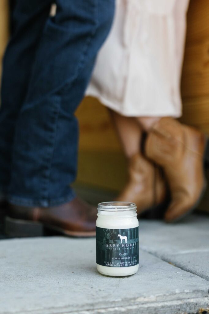 A Grey Horse Candle Co. jar candle sits on a concrete surface, with two people in boots standing close together and facing each other in the background, slightly out of focus.