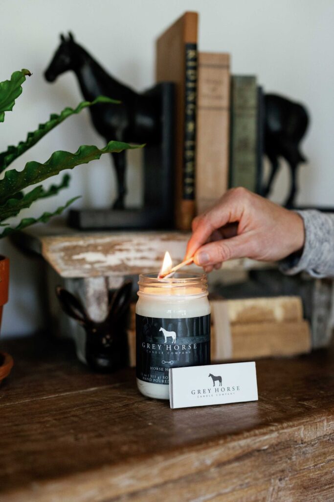 A hand lights a Grey Horse candle on a wooden table, with books, a potted plant, a horse figurine, and a Grey Horse matchbox in the background.